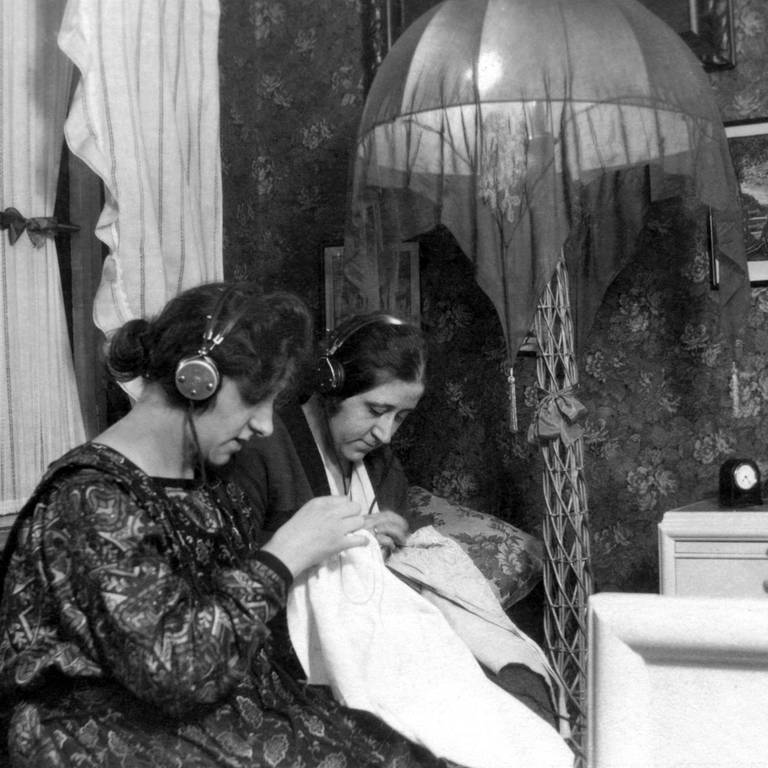 Zwei Frauen hören in den 1930ern mit Kopfhörer Radio und sticken: 1923 begann der reguläre Rundfunkbetrieb in Deutschland – maßgeblich vorangetrieben vom weitgereisten Ingenieur und Ministerialdirektor Hans Bredow