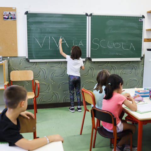 Klassenzimmer einer italienischen Grundschule. Ein kleines Mädchen schreibt auf die Tafel "viva la scuola", Es lebe die Schule.
