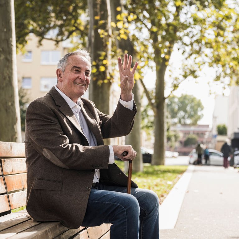 Ein lächelnder älterer Mann mit Spazierstock sitzt auf einer Bank und winkt.