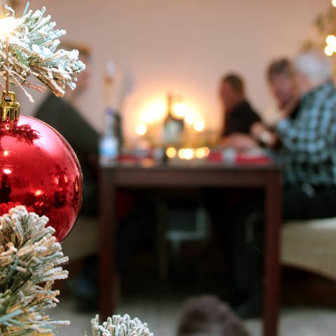 Im Vordergrund eine Weihnachtskugel die am Baum hängt, während leicht verschwommen im Hintergrund eine Familie Heiligabend am Tisch sitzt und isst.