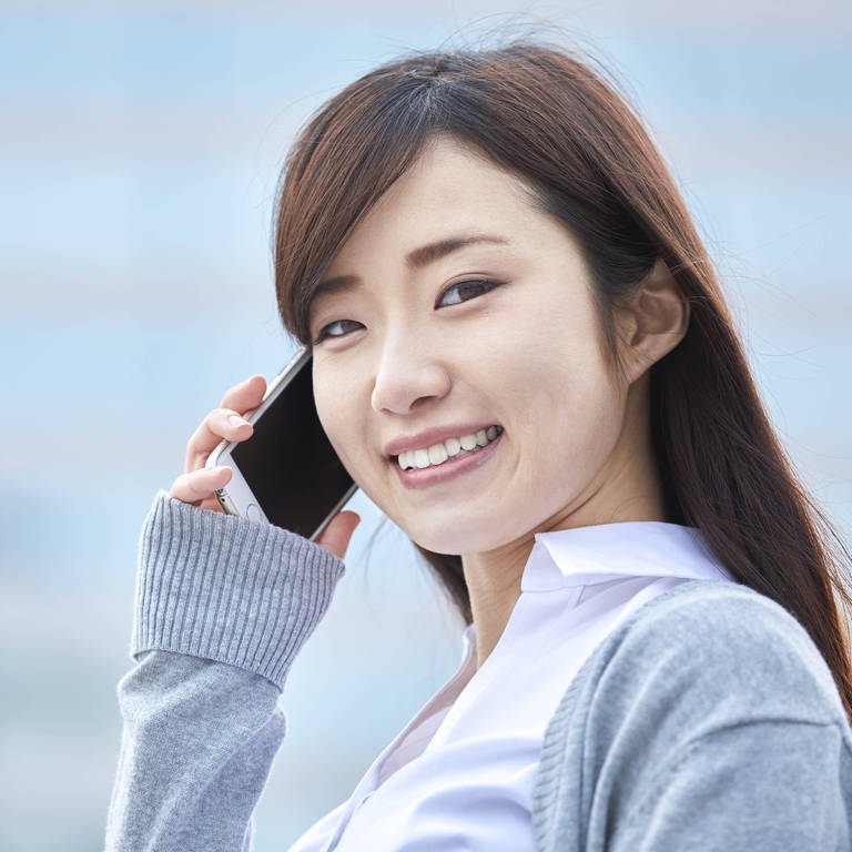 Junge japanische Geschäftsfrau mit Smartphone am Ohr lächelt: Bei der Gleichberechtigung gehört Japan zu den rückständigsten Staaten. Doch inzwischen wehren sich Japanerinnen mit wachsendem Erfolg gegen das tief verankerte Rollenbild der dienenden und dekorativen Frau. (Foto: IMAGO, IMAGO / AFLO)