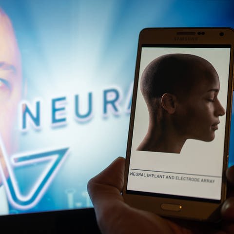 Neuralink-Illustration: Das Neuralink-Logo wird auf dem Mobilgerät angezeigt, im Hintergrund ist Gründer Elon Musk auf dem Bildschirm zu sehen