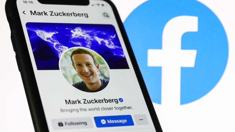 Das Facebook-Profil von Mark Zuckerberg, das auf einem Telefonbildschirm angezeigt wird, und das Facebook-Logo, das auf einem Bildschirm im Hintergrund angezeigt wird, sind auf diesem Illustrationsfoto zu sehen