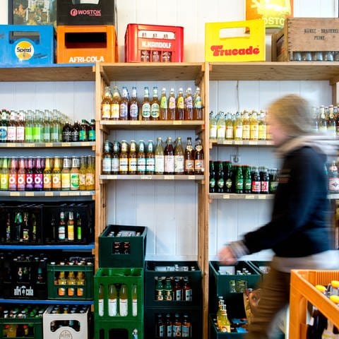 Eine Frau geht in einem Getränkemarkt an einem Regal vorbei, in dem Limonaden verschiedener Marken stehen.