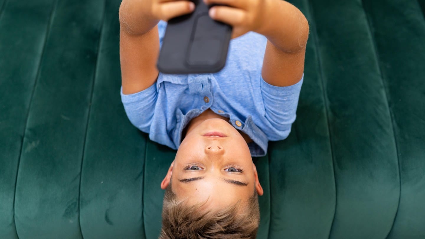 Blick aus der Vogelperspektive auf einen Jungen, der sein Mobiltelefon benutzt, während er auf dem Sofa im Wohnzimmer liegt (Foto: picture-alliance / Reportdienste, picture alliance / Zoonar | Channel Partners)