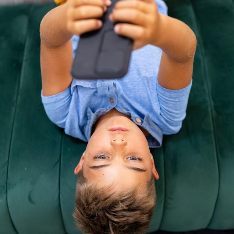 Blick aus der Vogelperspektive auf einen Jungen, der sein Mobiltelefon benutzt, während er auf dem Sofa im Wohnzimmer liegt