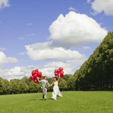 Braut und Bräutigam laufen mit roten Ballons über eine Wiese