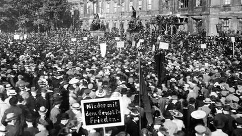 Bild einer Demonstration gegen die Vorgaben des Versailler Friedensvertrags in Berlin 1919,  Fahnen und Schilder, von denen eines "Nieder mit dem Gewaltfrieden" fordert, viele Hüte, schwarzweiß