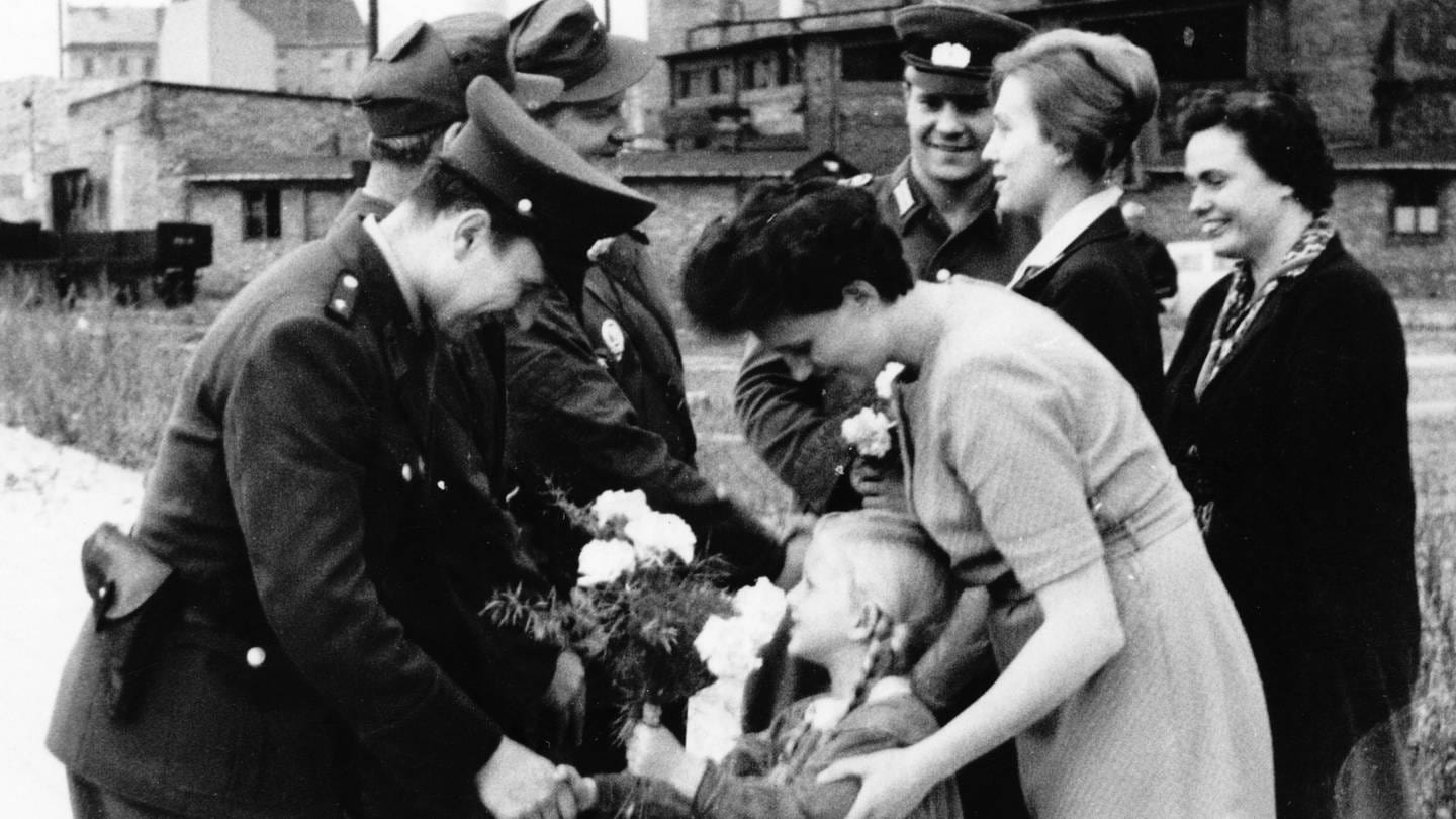 Ein Mädchen überreicht in Begleitung der Mutter Blumen an einen bewaffneten DDR-Grenzschützer. Original-Pressemeldung des DDR-Nachrichtendienstes ADN auf der Rückseite des Bildes: 