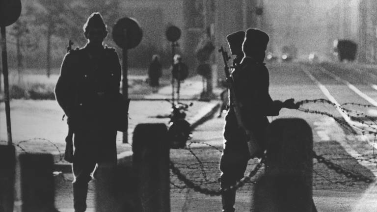Drei Volkspolizisten ziehen am Potsdamer Platz hinter einer Sperrkette eine Stacheldrahtbarriere. Im Hintergrund ist die menschenleere Leipziger Straße zu sehen. Am frühen Sonntagmorgen des 13. August 1961 wurde unter der Aufsicht von bewaffneten Streitkräften der DDR mit der Errichtung von Straßensperren aus Stacheldraht und dem Bau einer Mauer begonnen