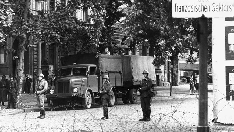 Hinter dem Stacheldrahtverhau in der Bernauer Straße stehen Volkspolizisten mit dem Gewehr im Anschlag. Am frühen Sonntagmorgen des 13. August 1961 wurde unter der Aufsicht von bewaffneten Streitkräften der DDR mit der Errichtung von Straßensperren aus Stacheldraht und dem Bau einer Mauer begonnen
