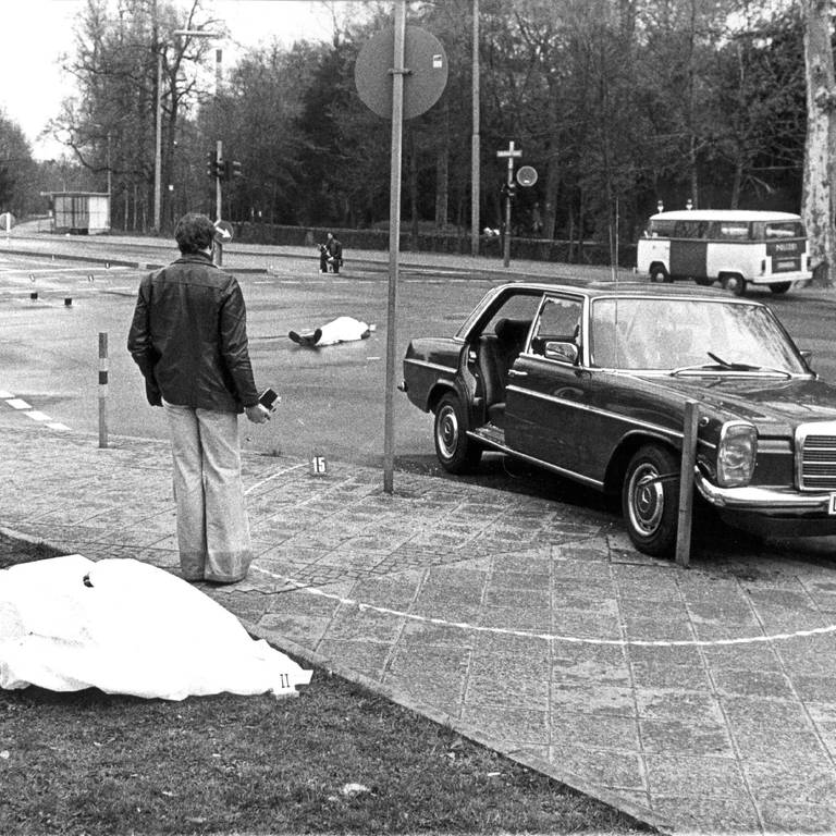 Der Tatort mit den zugedeckten Leichen von Siegfried Buback (vorne links) und seines Fahrers Wolfgang Göbel sowie der Dienstwagen des Generalbundesanwaltes, in dem die beiden erschossen wurde. Der höchste Ankläger der Bundesrepublik ist am 7. April 1977 in Karlsruhe durch Schüsse aus einer Maschinenpistole, die von einem Motorrad aus auf seinen Dienstwagen abgegeben worden waren, getötet worden. Mit ihm starb sein Fahrer Wolfgang Göbel, der lebensgefährlich verletzte Justizhauptwachtmeister Georg Wurster erlag am 13. April seinen Verletzungen. (Foto: dpa Bildfunk, picture-alliance / dpa/dpaweb | Heinz_Wieseler)