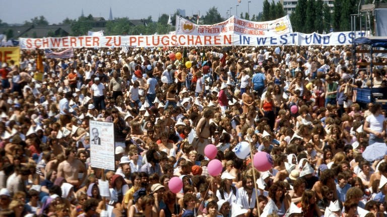 Demonstration auf den Rheinwiesen bei Bonn gegen die nukleare Nachrüstung am 10. Juni 1982. Etwa 500.000 Menschen nahmen teil. Im Hintergrund ein großes Transparent mit der Aufschrift: "Schwerter zu Pflugscharen – In West und Ost". Das Zitat "Schwerter zu Pflugscharen" hat seinen Ursprung in der Bibel und wurde auch in der Friedensbewegung zum geflügelten Wort. (Foto: IMAGO, IMAGO / Sven Simon)