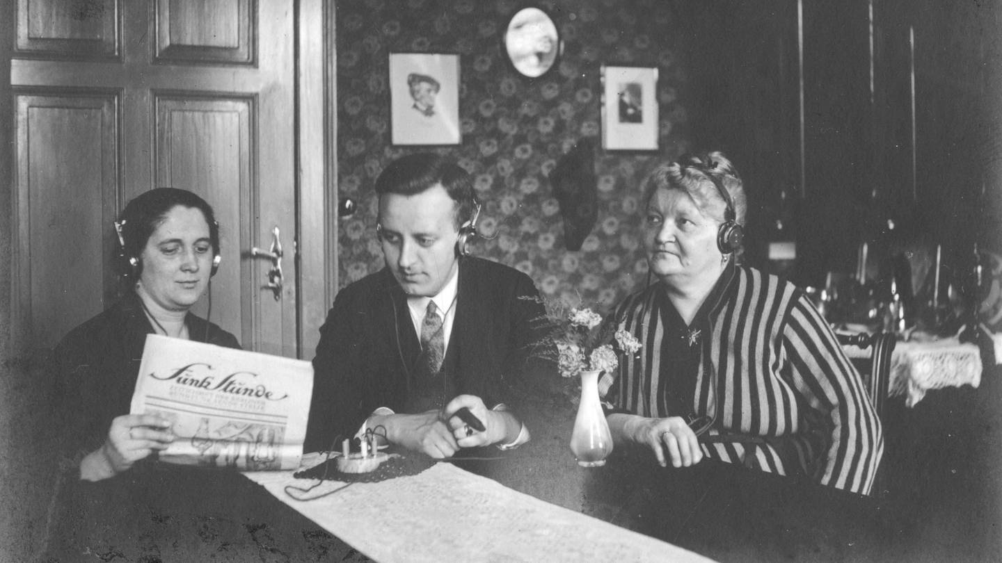 Drei Radiohörer, zwei Frauen, in der Mitte ein Mann, mit Kopfhörern und Detektorempfänger. Undatiertes Foto aus den 1920ern. Die Frau links im Bild hält ein Papier, vermutlich ein Programmheft, mit dem Titel 