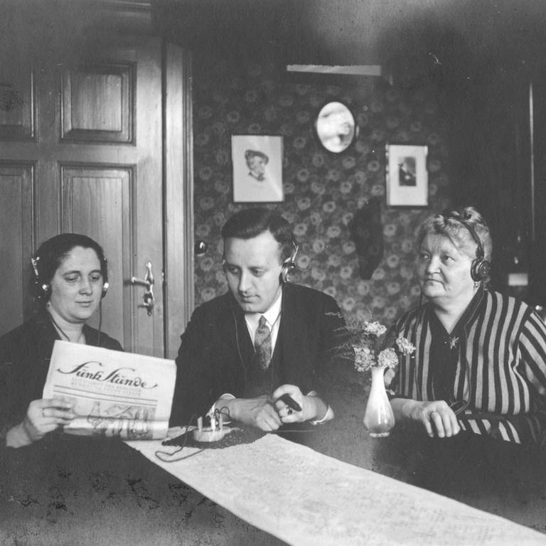 Drei Radiohörer, zwei Frauen, in der Mitte ein Mann, mit Kopfhörern und Detektorempfänger. Undatiertes Foto aus den 1920ern. Die Frau links im Bild hält ein Papier, vermutlich ein Programmheft, mit dem Titel "Funk Stunde" in der Hand.