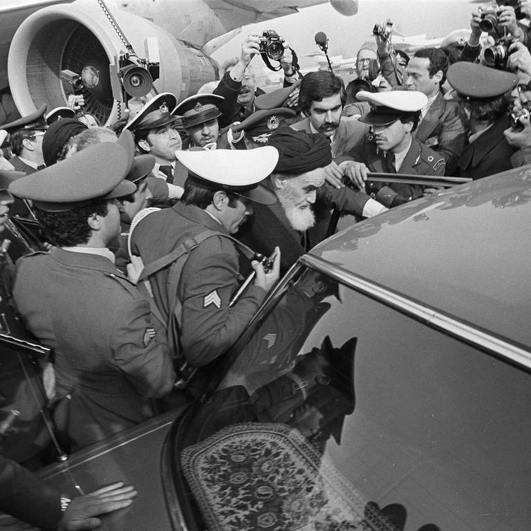 Ajatollah Khomeini wird stark bedrängt, als er am 1.2.1979 aus dem Pariser Exil in den Iran zurückkehrt. Sicherheitsleute schützen ihn, als er am Flughafen Teheran in ein Auto steigt.
