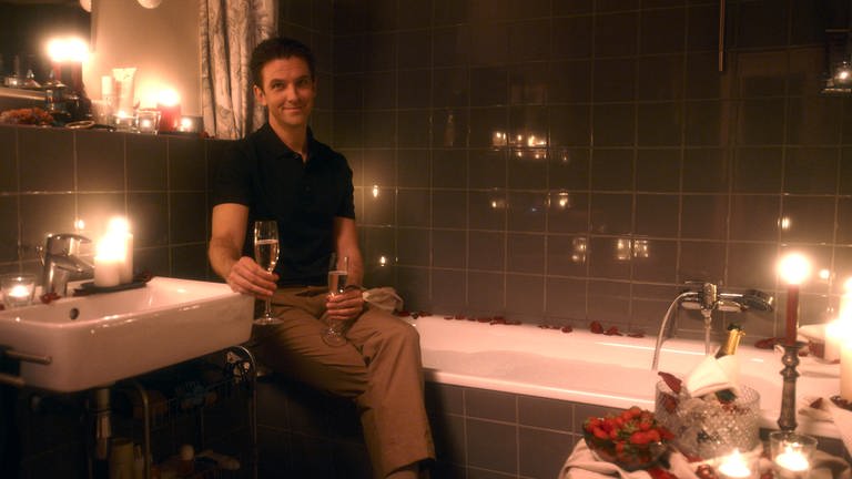 Dan Stevens sitzt auf dem Badewannenrand, zwei Gläser Champagner in den Händen, um ihn herum brennen Kerzen