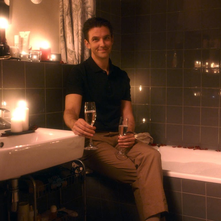 Dan Stevens sitzt auf dem Badewannenrand, zwei Gläser Champagner in den Händen, um ihn herum brennen Kerzen (Foto: SWR)