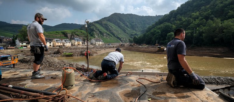Das Motiv zum Jahrestag der Flutkatastrophe in RLP zeigt drei Handwerker bei der Arbeit an der Ahr. Im Hintergrund fließt der Fluss und sind Weinberge zu sehen. (Foto: SWR, Harald Tittel, picture alliance/dpa)