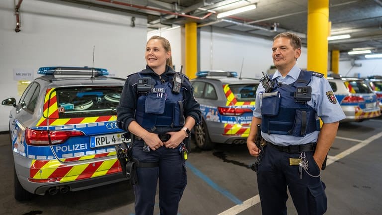 Katharina Klüpfel links) und Michael Kirch (rechts) stehen nach der Rückkehr vom Streifendienst in voller Polizeiausrüstung in der Tiefgarage vor Polizeiautos. SWR Dokuserie "Nachtstreife.