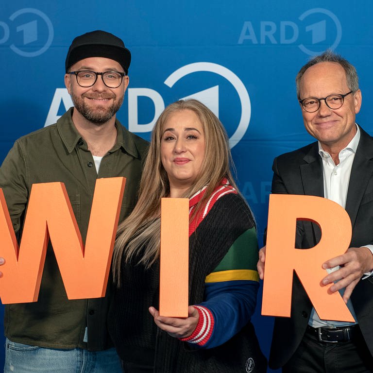 Musiker Mark Forster, Comedienne Idil Baydar und SWR Intendant Kai Gniffke (v.l.n.r.) mit orangenen Buchstaben "W I R" in den Händen, vor blauem ARD-Hintergrund