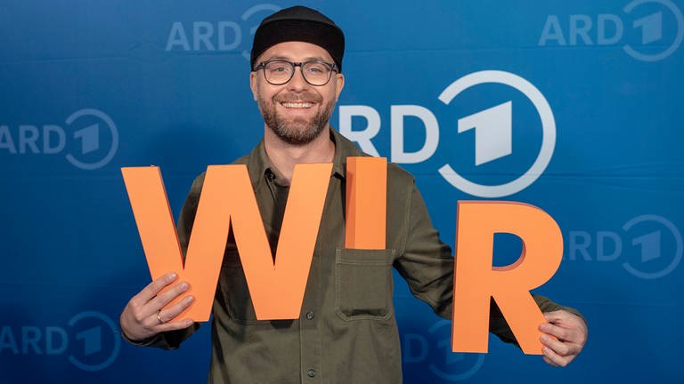 Mark Forster mit orangenen Buchstaben "W I R" vor blauem ARD-Hintergrund. (Foto: SWR, Thorsten Jander)