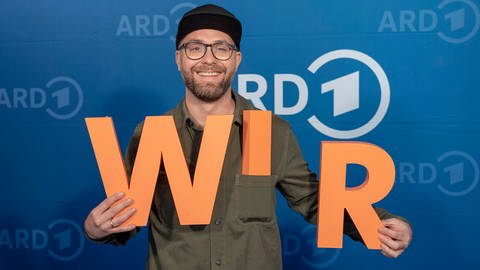 Mark Forster mit orangenen Buchstaben "W I R" vor blauem ARD-Hintergrund.