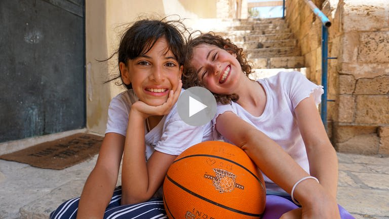 Schau in meine Welt. Judi (li.) ist Muslima und Lilya (re.) ist Jüdin, beide sind 11 Jahre alt und ein Herz und eine Seele. Sie sitzen fröhlich lachend auf dem Boden und lehnen an einen Basketball (Foto: SWR, Phare Media UG)