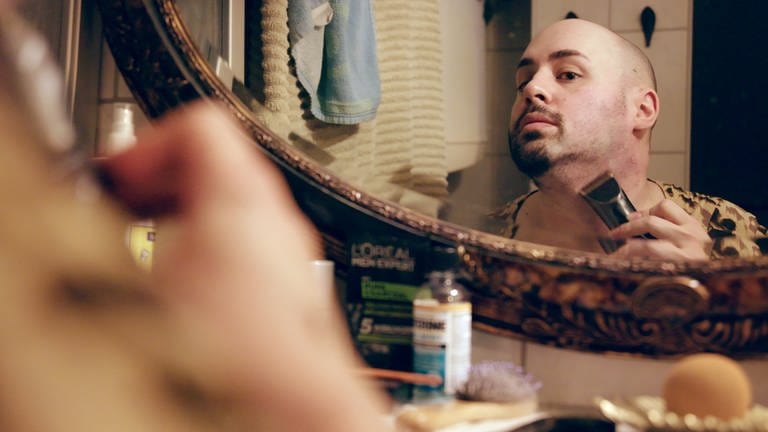 Jonas rasiert sich vor einem Spiegel seinen Bart, um in die Rolle von Dragqueen Macy M. Meyers zu schlüpfen.