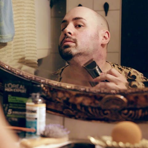 Jonas rasiert sich vor einem Spiegel seinen Bart, um in die Rolle von Dragqueen Macy M. Meyers zu schlüpfen.