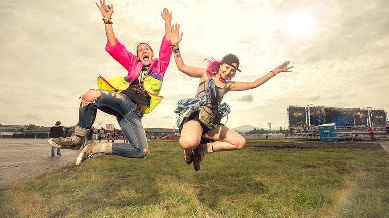 Zwei Menschen springen in die Luft und freuen sich scheinbar. Im Hintergrund erkennt man ein Festivalgelände.