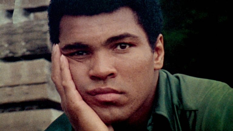 Boxer Muhammad Ali in einem dunkelgrünen Hemd sitzt vor einer Steinmauer, er stützt seinen Kopf in seine rechte Hand und blickt nachdenklich in die Kamera