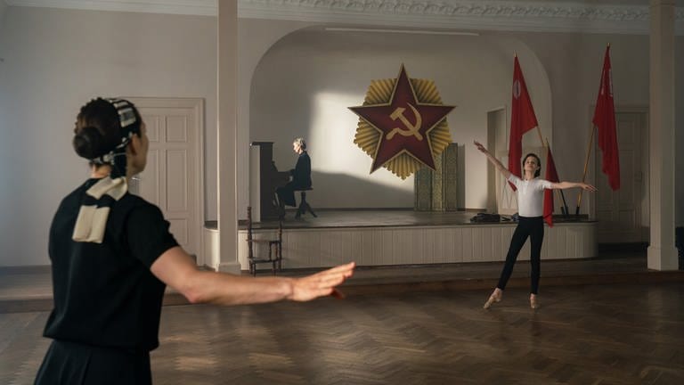 Rudolf (Cooper Dillon, re.) tanzt Ballett, seine Lehrerin steht links vorne im Bild und gibt ihm Anweisungen.