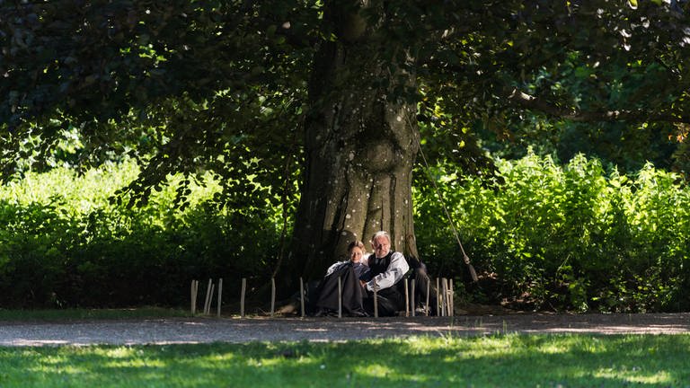 Mania (Nelly Hoffmann, li.) und ihr Vater (Max Herbrechter, re.) sitzen an einen Baum angelehnt und beobachten das schwingende Pendel. 
