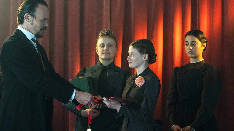 Mania (Nelly Hoffmann, mi.), so wird Marie Curie als Kind genannt, bekommt von Herrn Apuchtin (Robert Besta, li.) die Auszeichnung als Jahrgangsbeste überreicht. 