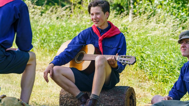 Herbert (Arthur Grupp) spielt Gitarre, auf einem Holzstamm sitzend.