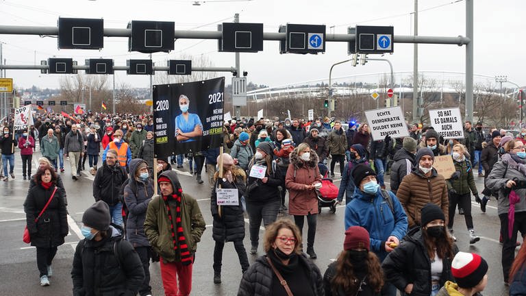 Viele gegen die Corona-Maßnahmen Protestierende laufen wegen der kalten Jahreszeit im Januar in dicke Jacken gehüllt und mit Mützen in einem Protestzug auf einer breiten Straße in Stuttgart. Einige tragen Schilder oder Plakate, manche mit, aber viele ohne Mund-Nasen-Schutz. (Foto: SWR)