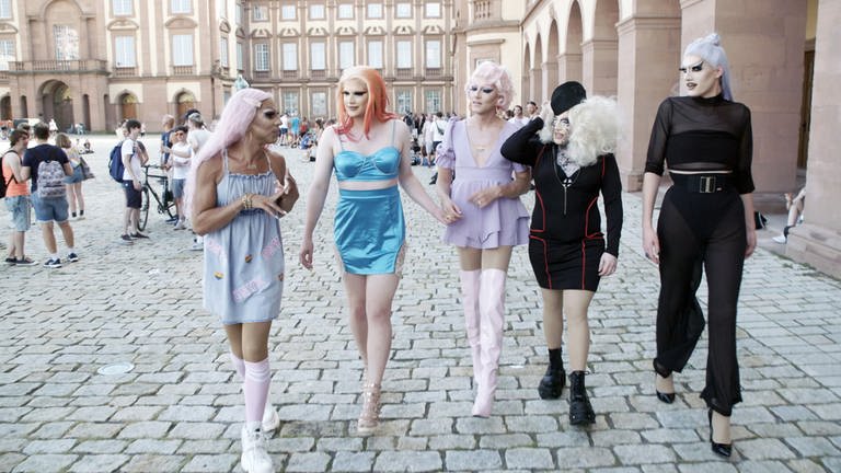 5 Dragqueens in glamouröser Aufmachung mit Perücken und Make-up flanieren in der Innenstadt von Mannheim. Bis auf die Person rechts tragen alle anderen kurze Kleider oder Röcke. (Foto: SWR)