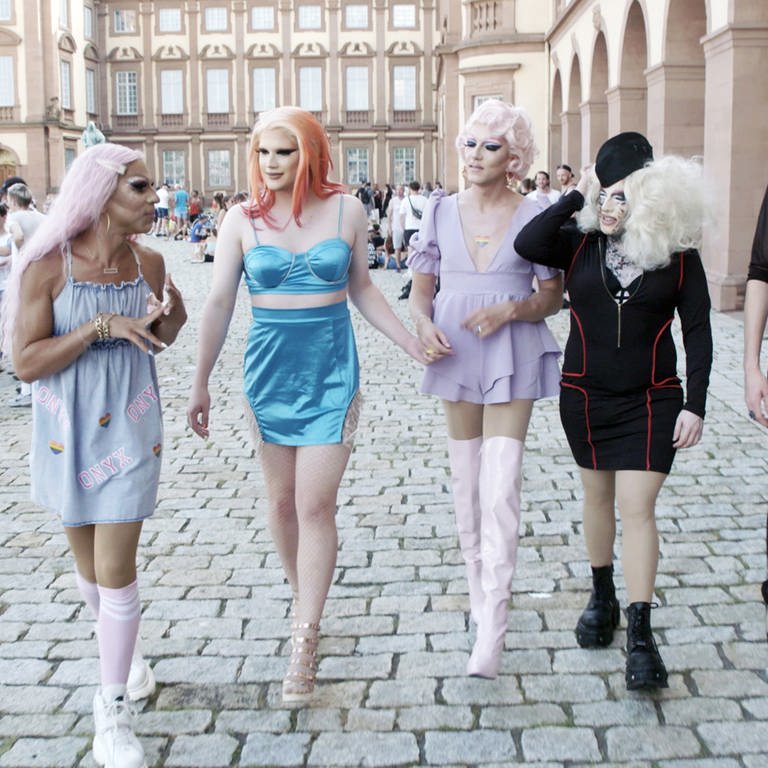 5 Dragqueens in glamouröser Aufmachung mit Perücken und Make-up flanieren in der Innenstadt von Mannheim. Bis auf die Person rechts tragen alle anderen kurze Kleider oder Röcke. (Foto: SWR)