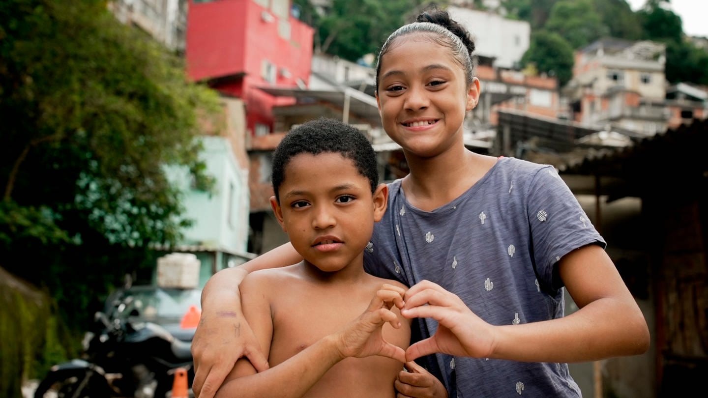Die Geschwister Maria-Eduarda (rechts) und Jose-Miguel leben in der Favela Rocinha. Sie formen gemeinsam mit den Fingern ein Herz. Im Hintergrund sind Häuser des Armenviertels zu sehen. Doku „Tür an Tür mit dem Gangster – Leben in Rios größter Favela“ in der ARD Mediathek. (Foto: SWR)