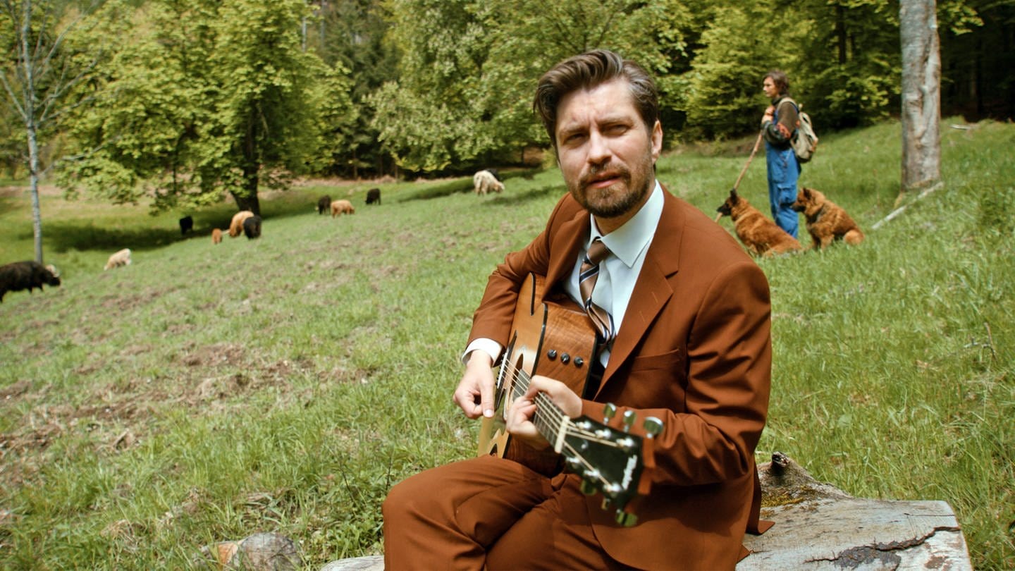 David Julian Kirchner spielt ein Lied auf der Gitarre inmitten einer Kuh-Herde (Foto: SWR, Donni Schönemond)