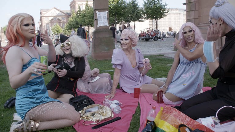 Die fünf Protagonisten der Doku sitzen bei schönem Wetter auf zwei rosa-farbenen Picknick-Decken auf einem Rasenstück vor einem Platz und trinken, lachen, unterhalten sich lebhaft. Alle tragen ihre Kostüme, Perücken und Make-up. (Foto: SWR)
