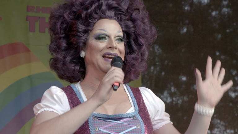 Die bekannte Mannheimer Dragqueen Celine Bouvier trägt eine üppige aubergine-farbene Perücke und eine farblich passendes Dirndl. Sie hält ein Mikrofon und ist auf einer Open-Air-Bühne, im Hintergrund ein Plakat einem Regenbogen, dem Symbol der Lesben- und Schwulenbewegung.