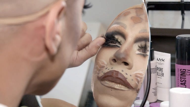 Blick über die Schulter in einen Spiegel zeigt die Dragqueen Miss Onyx beim Styling mit falschen Wimpern und viel kunstvollem Make-up.