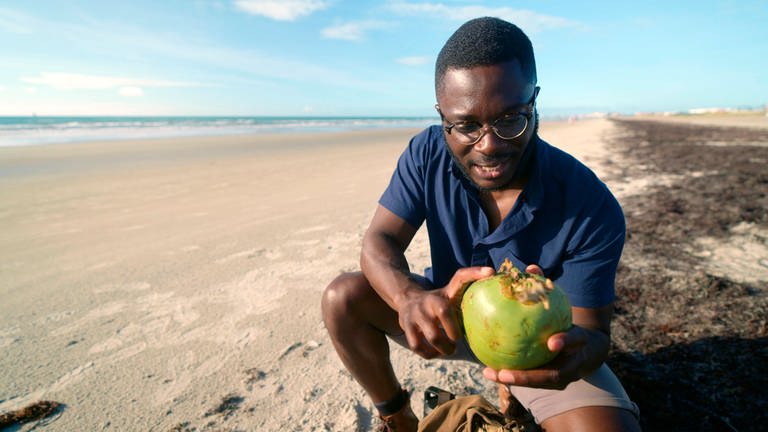 Patrick Aryee, BBC-Wildlifeexperte und Dokumentarfilmer, an einem Strand. Er hält eine grüne Frucht in den Händen und erklärt deren Besonderheiten. (Foto: SWR, SHE/Beach House Pictures)