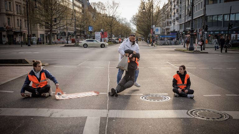 ARD Story: Radikal fürs Klima - Helden oder Kriminelle? Aktivisten der "Letzten Generation" werden bei einer Straßenblockade auf dem Kurfürstendamm von Beamten von der Straße weggezerrt. (Foto: SWR, © SWR/Gordon Welters)