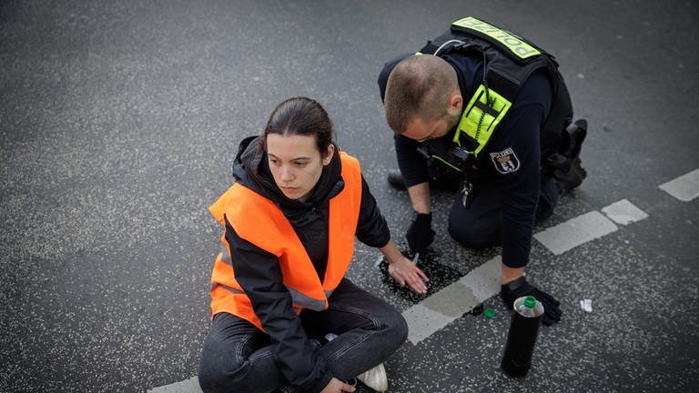 "ARD Story: Radikal fürs Klima - Helden oder Kriminelle?" Ein Polizist löst die angeklebte Hand einer Aktivistin bei einer Straßenblockade der "Letzten Generation" auf dem Kurfürstendamm in Berlin. (Foto: SWR, © SWR/Gordon Welters)
