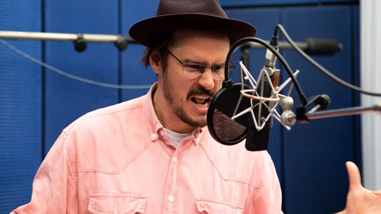 Tilman Strauß, mit Bart und Brille, gekleidet mit rosa Hemd und braunem Hut, steht im Hörspiel-Studio vor einem Mikrofon. Er spricht im Hörspiel "Der Termin" "Der Termin", nach dem gleichnamigen Roman von Katharina Volckmer.