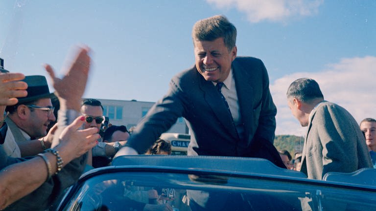 Senator John F. Kennedy sitzt in einem Cabrio und begrüßt die Menschenmenge am Straßenrand