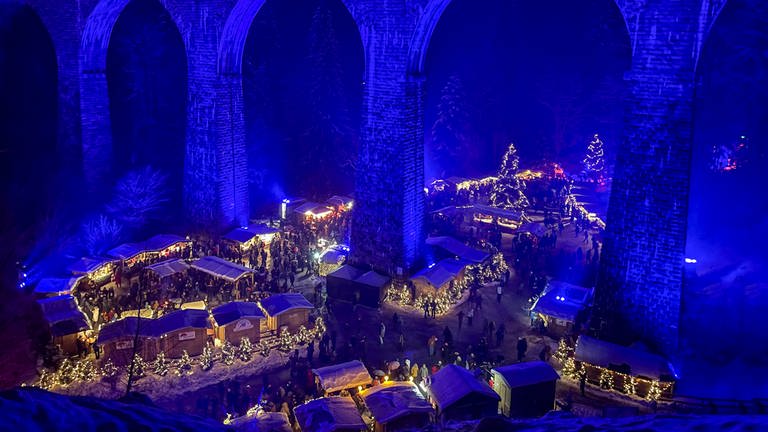Beleuchtete Weihnachtsmarkstände stehen in der Ravennaschlucht, darüber verläuft die in blaues Licht getauchte Ravennabrücke